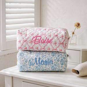 May include: Two rectangular quilted pouches stacked vertically. The top pouch is white with a pink floral and ribbon pattern, featuring the name Eloise embroidered in pink. The bottom pouch is white with a blue floral pattern, featuring the name Maria embroidered in blue.