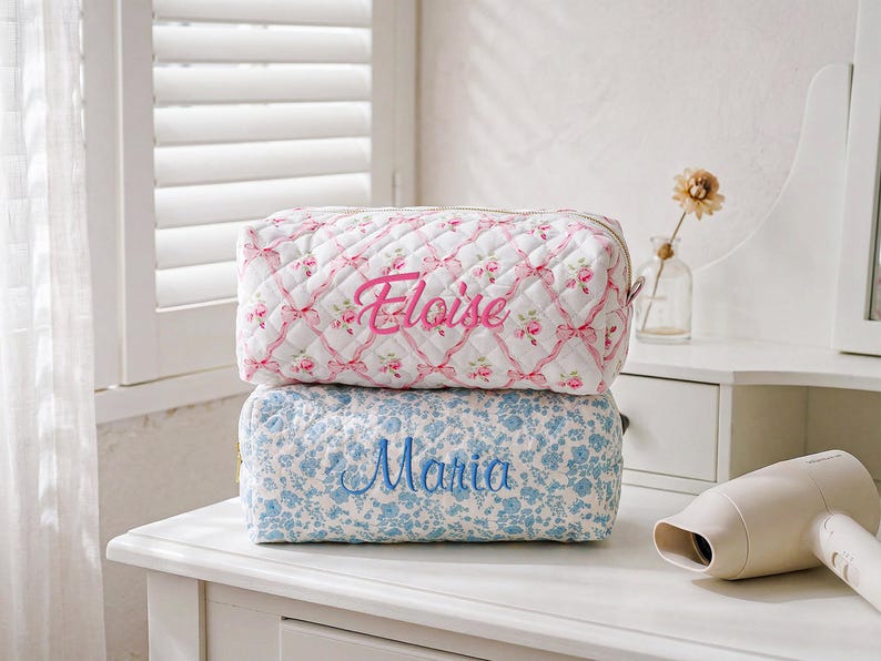 May include: Two rectangular quilted pouches stacked vertically. The top pouch is white with a pink floral and ribbon pattern, featuring the name Eloise embroidered in pink. The bottom pouch is white with a blue floral pattern, featuring the name Maria embroidered in blue.