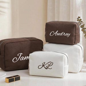 Puede incluir: Cuatro neceseres personalizados en marrón y blanco. Las bolsas marrones tienen los nombres "Jam" y "Audrey" bordados en escritura blanca. Las bolsas blancas tienen las iniciales "KL" bordadas en escritura negra. Todas las bolsas tienen una textura de gofre.