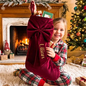 May include: A deep red velvet Christmas stocking featuring a large, prominent bow on the front. The name Lilian is embroidered in gold script on one of the hanging tails of the bow. The stocking has a loop at the top for hanging purposes.