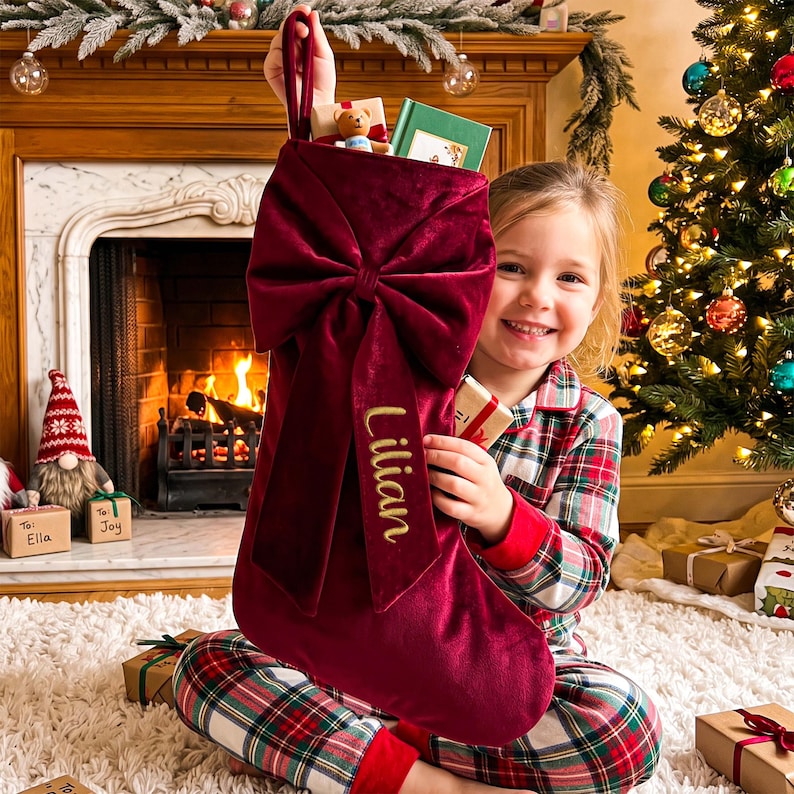 May include: A deep red velvet Christmas stocking featuring a large, prominent bow on the front. The name Lilian is embroidered in gold script on one of the hanging tails of the bow. The stocking has a loop at the top for hanging purposes.