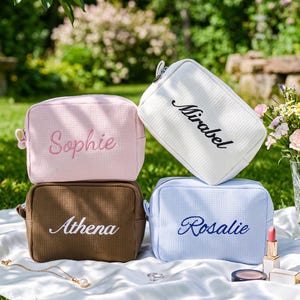 Pu&ograve; includere: Quattro trousse per cosmetici personalizzate in rosa, bianco, marrone e blu, ciascuna ricamata con un nome: Sophie, Mirabel, Athena e Rosalie. Le borse sono disposte su un tessuto bianco con trucco e gioielli nelle vicinanze.