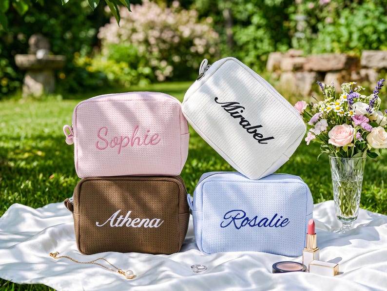 Pu&ograve; includere: Quattro trousse per cosmetici personalizzate in rosa, bianco, marrone e blu, ciascuna ricamata con un nome: Sophie, Mirabel, Athena e Rosalie. Le borse sono disposte su un tessuto bianco con trucco e gioielli nelle vicinanze.
