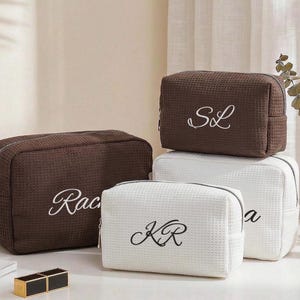 May include: Three rectangular cosmetic bags with a waffle-textured fabric. One large dark brown bag features the name Rachel, a smaller dark brown bag is embroidered with SL, and a small white bag displays the monogram KR in black embroidery.