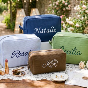 Pu&ograve; includere: Quattro trousse per cosmetici personalizzate in blu, azzurro, verde e marrone, ciascuna ricamata con un nome o iniziali in un colore a contrasto. Le trousse sono esposte su una tovaglia di pizzo con trucco e gioielli.