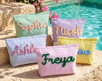 May include: Five seersucker cosmetic bags in various colors: green, purple, blue, pink, and yellow. Each bag has a name embroidered on it in a contrasting color. The bags are displayed near a pool.