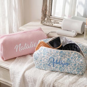 May include: A pink waffle-textured toiletry bag with the name Natalie embroidered in white, and a blue floral-patterned quilted toiletry bag with the name Addison embroidered in blue, both featuring zippered closures and rectangular shapes.