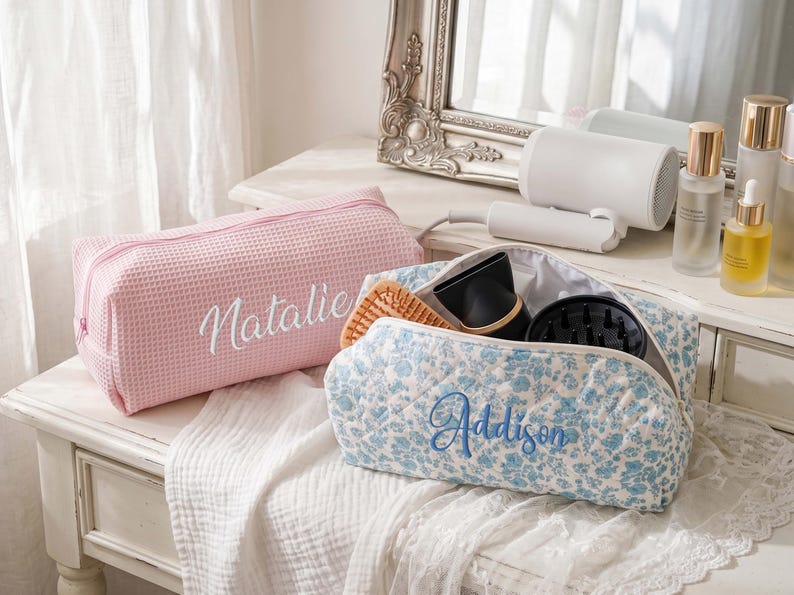 May include: A pink waffle-textured toiletry bag with the name Natalie embroidered in white, and a blue floral-patterned quilted toiletry bag with the name Addison embroidered in blue, both featuring zippered closures and rectangular shapes.