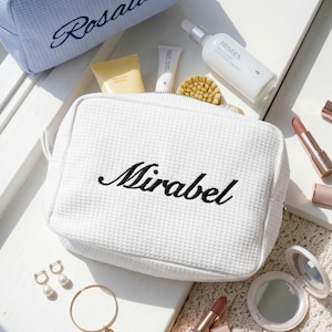 Pu&ograve; includere: Borse cosmetiche bianche e azzurre con texture a nido d'ape e i nomi ricamati "Mirabel" e "Rosalie". Le borse sono circondate da prodotti di bellezza, tra cui rossetti, un specchietto compatto e prodotti per la cura della pelle. Sono presenti anche gioielli in oro, tra cui orecchini e un braccialetto.