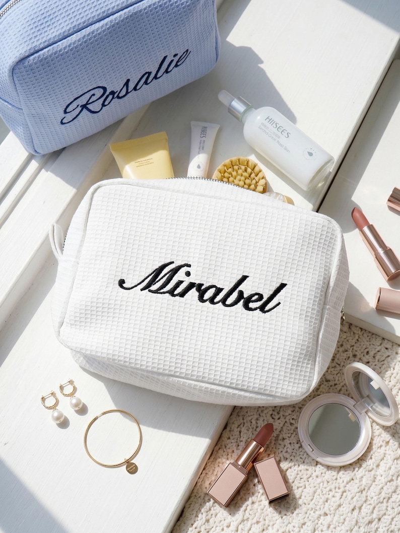 Pu&ograve; includere: Borse cosmetiche bianche e azzurre con texture a nido d'ape e i nomi ricamati "Mirabel" e "Rosalie". Le borse sono circondate da prodotti di bellezza, tra cui rossetti, un specchietto compatto e prodotti per la cura della pelle. Sono presenti anche gioielli in oro, tra cui orecchini e un braccialetto.