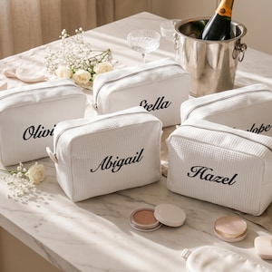 May include: Five white rectangular cosmetic bags with a waffle-textured fabric and a zip closure. Each bag features a custom name embroidered in black script lettering, specifically the names Olivia, Stella, Abigail, Penelope, and Hazel.