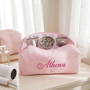 May include: A light pink waffle-textured fabric travel bag with the name Athena embroidered in dark pink script on the front. The open bag contains three hair dryer attachments, including a diffuser, and is positioned near a white hair dryer.