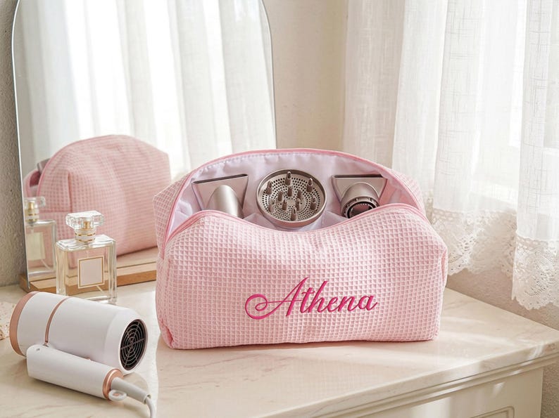 May include: A light pink waffle-textured fabric travel bag with the name Athena embroidered in dark pink script on the front. The open bag contains three hair dryer attachments, including a diffuser, and is positioned near a white hair dryer.