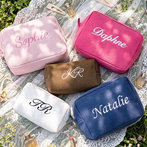 Pu&ograve; includere: Cinque trousse per cosmetici personalizzate in rosa, fucsia, marrone, bianco e blu navy, ciascuna ricamata con un nome o iniziali in un colore a contrasto. Le borse rettangolari hanno una superficie strutturata e una chiusura a zip. Sono esposte su un panno di pizzo bianco.