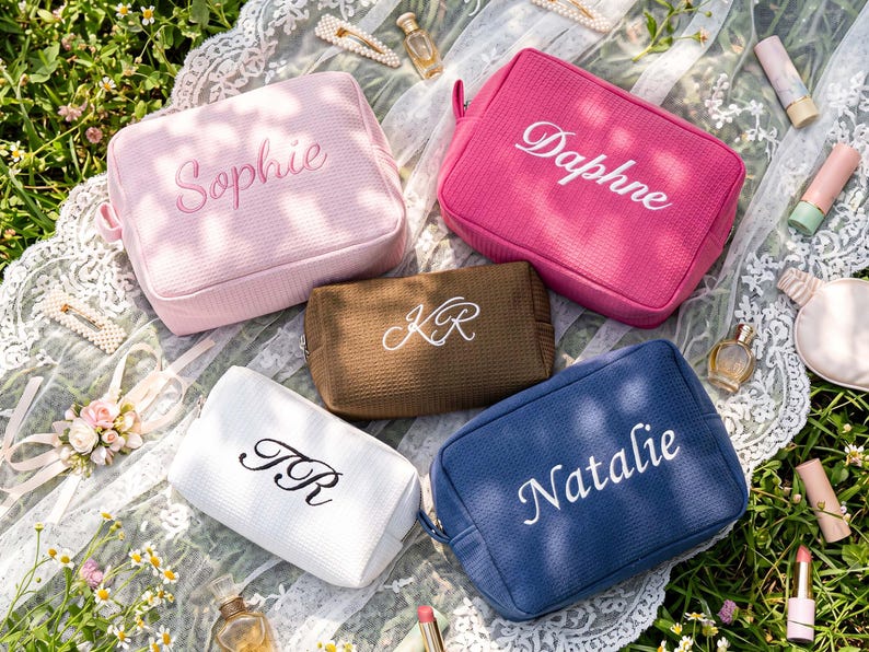 Pu&ograve; includere: Cinque trousse per cosmetici personalizzate in rosa, fucsia, marrone, bianco e blu navy, ciascuna ricamata con un nome o iniziali in un colore a contrasto. Le borse rettangolari hanno una superficie strutturata e una chiusura a zip. Sono esposte su un panno di pizzo bianco.
