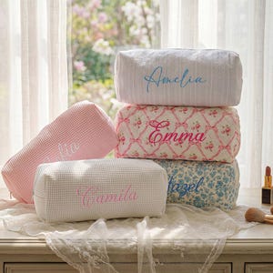 May include: A collection of rectangular cosmetic bags featuring various textures and patterns. One white bag is embroidered with the name Amelia, a floral print bag with Emma, a pink waffle-knit bag, a white waffle-knit bag with Camila, and a blue floral bag.