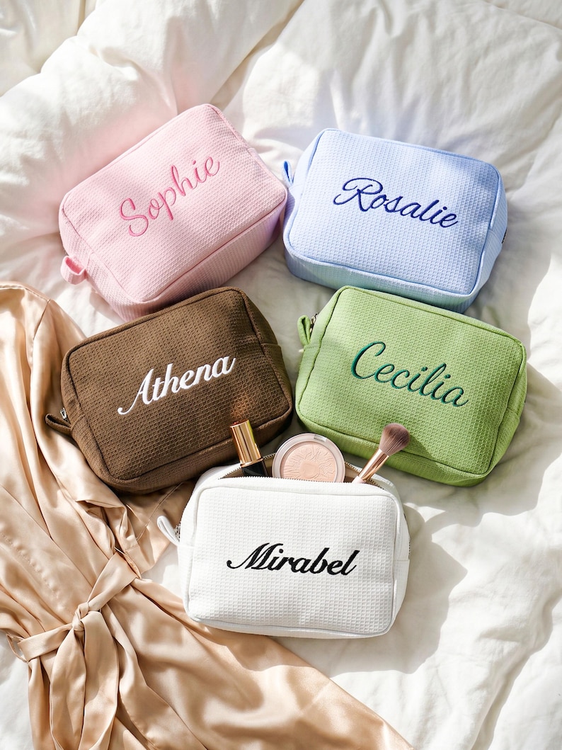 Pu&ograve; includere: Cinque trousse per cosmetici personalizzate in rosa, azzurro, marrone, verde e bianco. Ogni borsa presenta un nome diverso ricamato in un colore coordinato. La borsa bianca &egrave; aperta, mostrando trucchi.