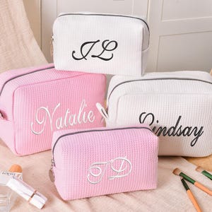 Peut inclure: Quatre trousses de maquillage personnalisées blanches et roses, avec des noms et initiales brodés en noir. Les sacs ont un motif gaufré texturé et des fermetures éclair noires. Des pinceaux de maquillage et un flacon de parfum sont au premier plan.