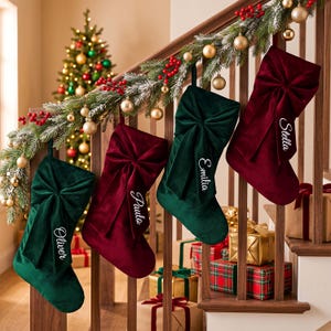 May include: Four velvet Christmas stockings in alternating dark green and deep red colours, each featuring a large decorative bow and personalised with white embroidered names: Oliver, Paula, Emilia, and Stella, hanging from a wooden staircase.