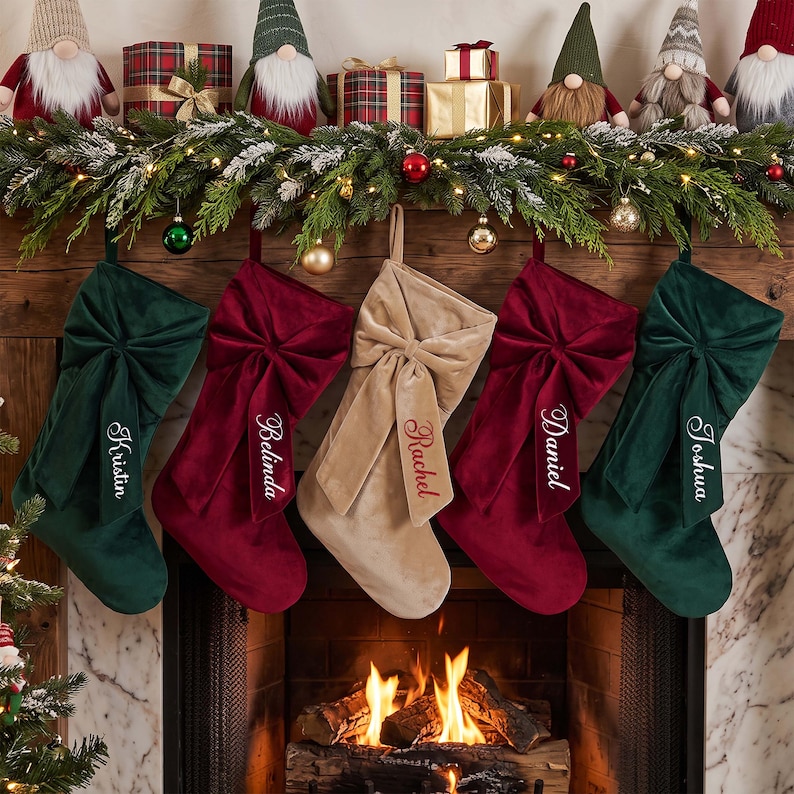 May include: Five velvet Christmas stockings hanging in a row, featuring two dark green, two deep red, and one beige stocking. Each stocking has a large decorative bow and is embroidered with a name: Kristin, Belinda, Rachel, Daniel, and Joshua.