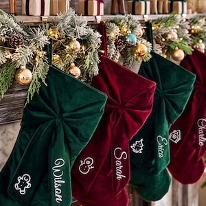 May include: A row of velvet Christmas stockings in deep green and burgundy, each featuring a large central bow and white embroidered names including Wilson, Sarah, and Erica, alongside small icons of a gingerbread man, snowman, and tree.