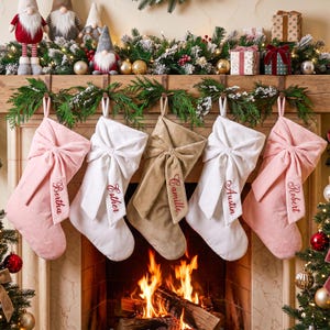 May include: Five personalized Christmas stockings hanging from a mantel, featuring large fabric bows and embroidered names: Bertha, Esther, Camille, Austin, and Robert. The stockings are in shades of soft pink, white, and tan velvet fabric.