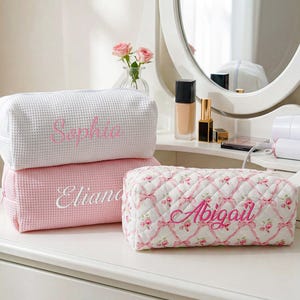 May include: Three rectangular cosmetic bags with zipper closures. One white waffle-textured bag features the name Sophia in pink embroidery. One pink waffle-textured bag features the name Eliana in white. One quilted bag with a pink floral ribbon pattern features the name Abigail in pink.