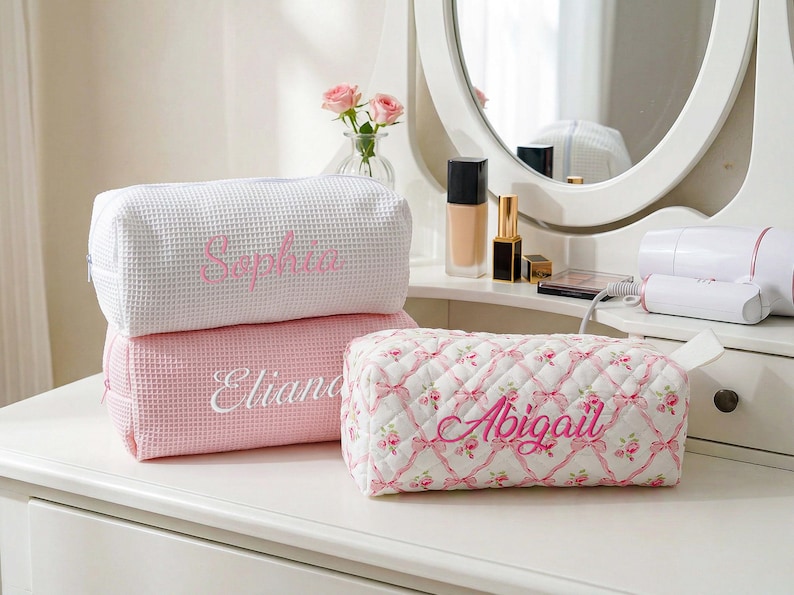 May include: Three rectangular cosmetic bags with zipper closures. One white waffle-textured bag features the name Sophia in pink embroidery. One pink waffle-textured bag features the name Eliana in white. One quilted bag with a pink floral ribbon pattern features the name Abigail in pink.