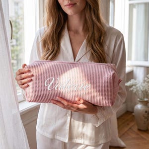 May include: A rectangular, light pink waffle-textured toiletry bag featuring a zipper closure along the top. The front of the bag is embroidered with the name "Valerie" in white cursive script lettering, centered on the textured fabric surface.