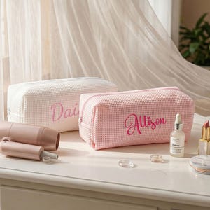 May include: Two rectangular waffle-weave cosmetic bags, one white and one light pink, each featuring a zippered closure and custom embroidery in pink script. The white bag displays the name Daisy, while the pink bag displays the name Allison.