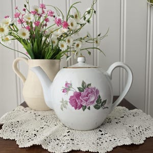 Può includere: Una teiera in ceramica bianca con un motivo floreale di rose rosa e verdi. La teiera è appoggiata su un centrino di pizzo color crema. Sullo sfondo c'è un brocca color crema con fiori bianchi e rosa.