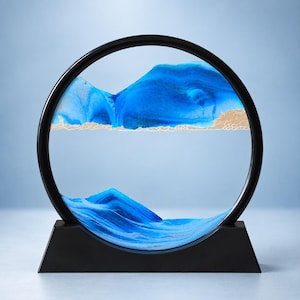 May include: A circular black frame containing a dynamic sand art display. The interior features layers of vibrant blue sand forming mountain-like peaks, contrasted by a horizontal band of textured gold sand that separates the upper and lower sections.