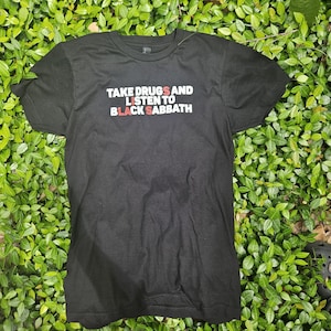 May include: Black t-shirt with the text "TAKE DRUGS AND LISTEN TO BLACK SABBATH" in white and red lettering. The shirt is made of a soft material and has short sleeves.