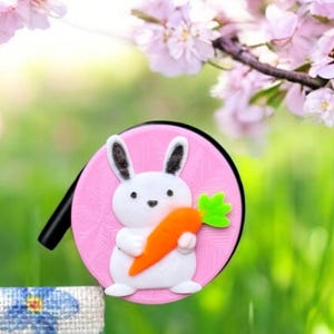 May include: A circular pink accessory featuring a white bunny holding a carrot, set against a backdrop of pink blossom and green grass. A floral patterned fabric is visible. The scene evokes springtime.
