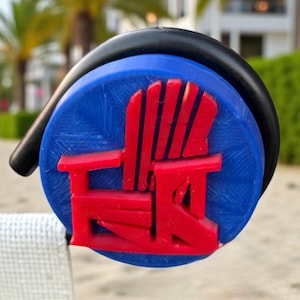 May include: A blue and red beach chair logo on a round, black-handled item. The logo is a red chair design on a blue background. The item is attached to a white and red striped beach towel on a sandy beach.