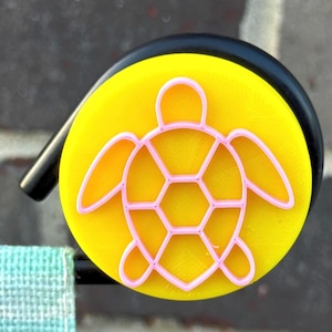 May include: A yellow disc with a pink turtle design. The turtle outline is pink, set against a yellow backdrop. A black handle is attached, alongside a light blue and green fabric.