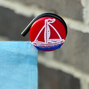 May include: A red, circular clip with a black handle. The clip features a white sailboat design with blue waves on a red background. The clip is attached to a light blue fabric, with a brick wall in the background.