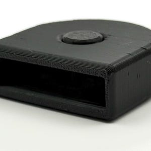 May include: A black, rectangular object with a rounded top and a circular feature. The object has a hollowed-out section on one side. The item is made of a textured material and sits on a white surface.