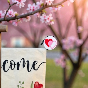 May include: A garden flag with the word "come" in black script, decorated with red heart and floral motifs. A small charm with two overlapping hearts in red and pink is attached. The background features pink blossom-covered branches.