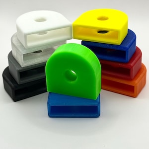 Puede incluir: Una colección de hebillas de plástico semicirculares de colores, incluyendo blanco, amarillo, azul, rojo, naranja, verde, gris y negro. Cada hebilla tiene una abertura rectangular y un agujero circular.