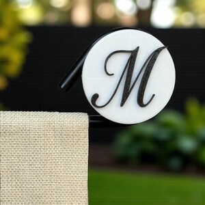 May include: A decorative towel holder featuring a white circular design with a black cursive letter "M". The holder has a black curved arm and is mounted on a black bar. A beige towel hangs below.