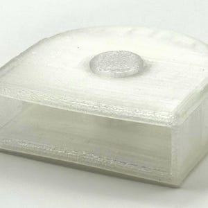 May include: A translucent, rectangular container with a rounded top and a small, circular, textured detail on top. The object appears to be made of a semi-transparent material, possibly plastic, and is designed with a hollow interior.