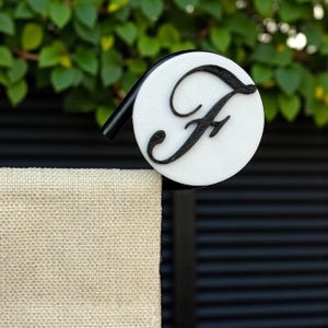 May include: A white and black bottle stopper with the letter "F" in a cursive font. The stopper has a circular design with a black stem. The background features green foliage and a textured surface.