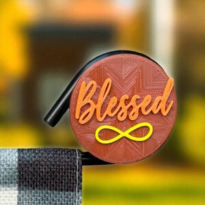May include: A brown, circular ornament with the word "Blessed" in orange script and a yellow infinity symbol. The ornament has a textured surface and a black curved handle. The background is blurred.