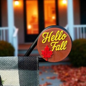 Może przedstawiać: Okrągły, brązowy znak z napisem "Hello Fall" w żółtym skrypcie i czerwonym liściem klonu. Znak jest przymocowany do czarnej metalowej ramy, z czarno-białą tkaniną w kratę na pierwszym planie.