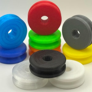 May include: A selection of multi-coloured 3D-printed spools. The spools are in shades of blue, red, green, grey, yellow, black, and white. Each spool has a central hole and a grooved design, stacked in small groups.