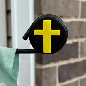 May include: A black and yellow cross-shaped flag holder. The holder is round with a yellow cross on a black background. The holder is attached to a black rod, and a light green flag with white daisies is visible.