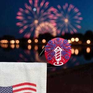 May include: A red, white, and blue firework-themed bottle stopper with a black handle. The stopper features a firework design with stars. The background shows fireworks over a body of water.