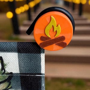 May include: A decorative orange and yellow campfire design. The circular design features a yellow flame and brown logs. The design is attached to a black, curved object. The background is blurred, with a black and white checkered pattern in the foreground.