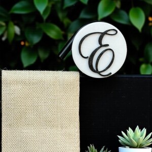 May include: A white and black whistle with a large, stylized letter "E" on the front. The whistle has a black mouthpiece and is set against a backdrop of green foliage and a black surface.